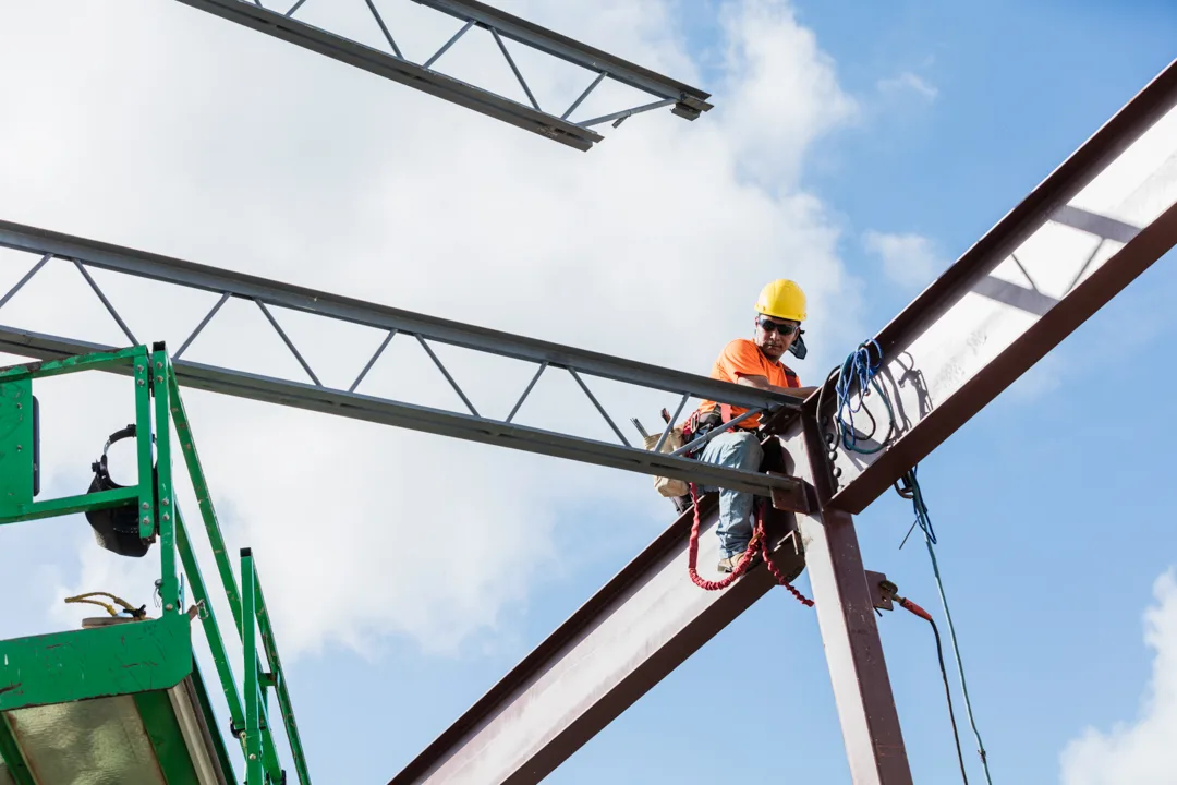 A Hispanic steel worker working high up on a girder. He is sitting on the girder, wearing a safety harness, working to secure a roof joist or truss to the girder.